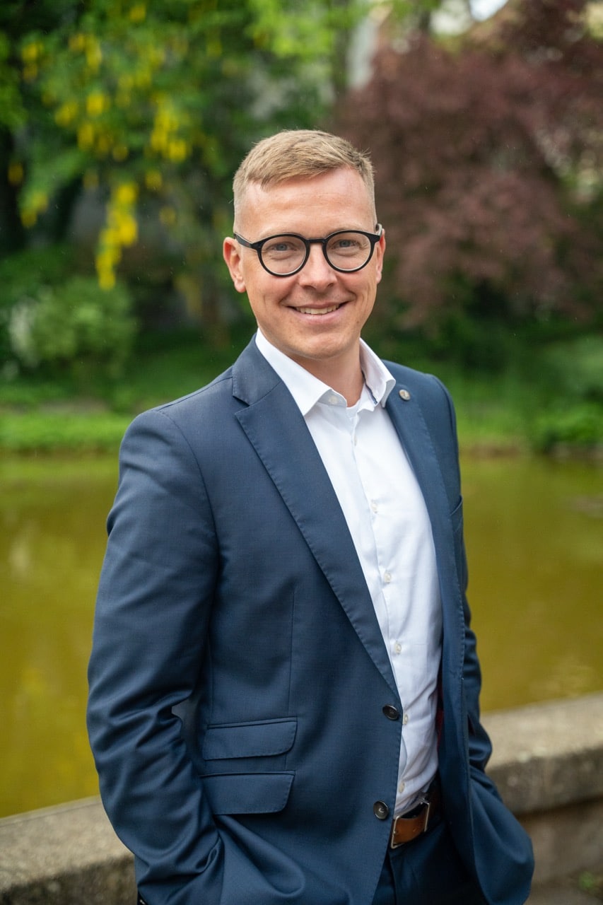 Kapitalkongress Kongresspaket – Videokongress zur Geldanlage 3 Portraitfoto von Florian Günther in dunklem Businessblazer und Brille vor grünem Garten