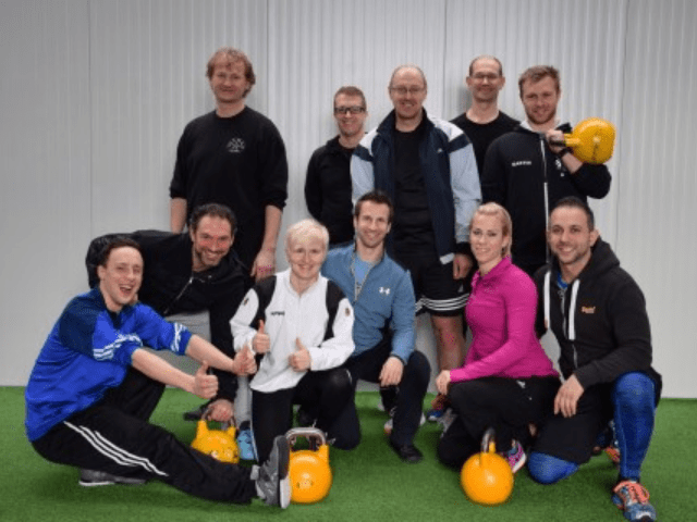 Etwa 14 Personen mit gelben Kettlebells auf grüner Trainingsmatte, Gruppenfoto