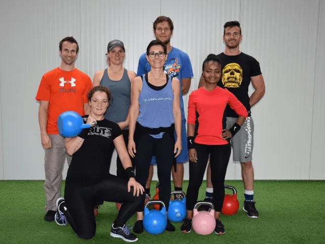Sechs Personen mit bunten Kettlebells auf grüner Trainingsmatte, Gruppenfoto