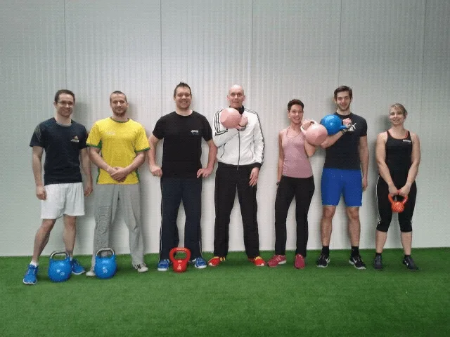 Sieben Personen mit verschiedenfarbigen Kettlebells auf grüner Trainingsmatte, Gruppenfoto