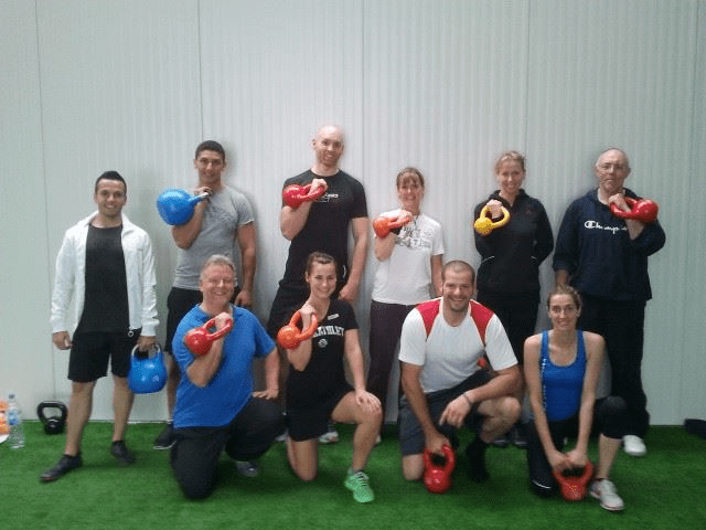 Gruppenfoto von etwa 12 Personen mit Kettlebells auf grünem Kunstrasen vor weißer Wand