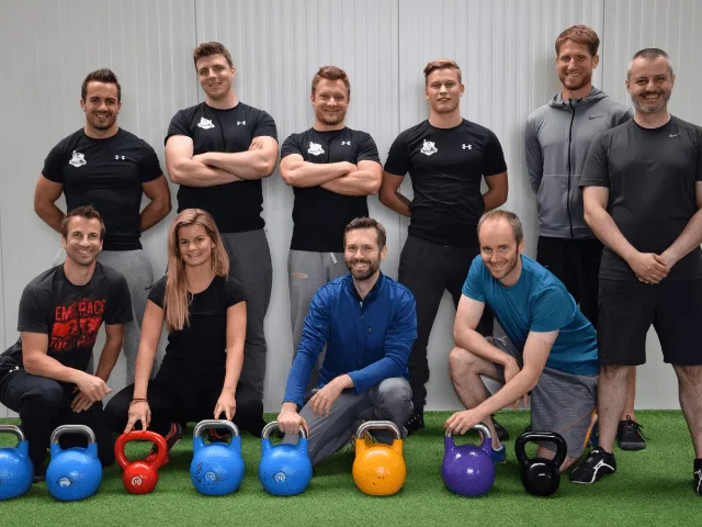 Zehn Personen in Trainingskleidung mit bunten Kettlebells auf grüner Trainingsmatte