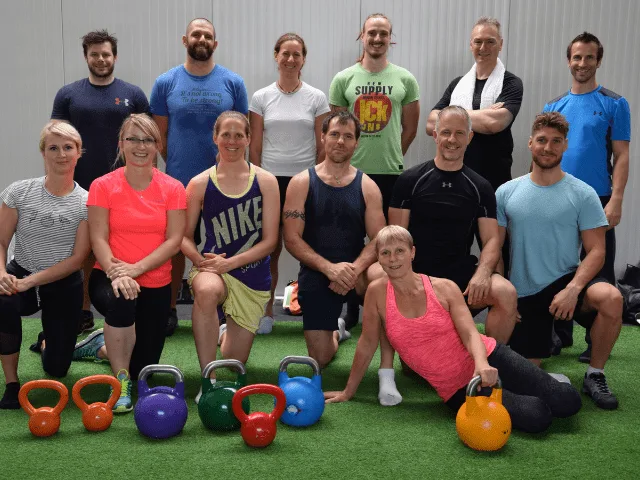 Gruppe von etwa 18 Personen mit verschiedenfarbigen Kettlebells auf grüner Trainingsmatte