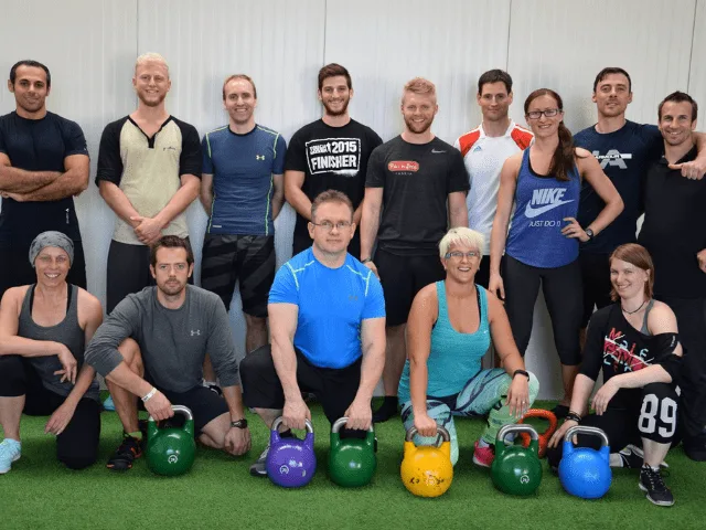 Etwa 20 Personen unterschiedlichen Alters mit Kettlebells auf grüner Trainingsmatte