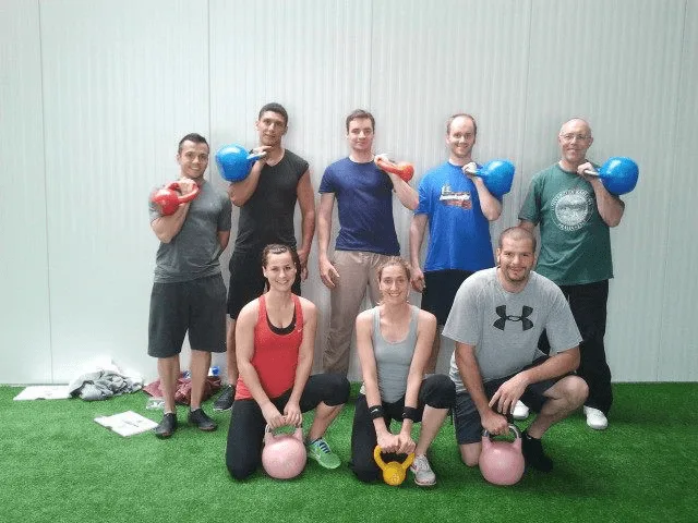 Neun Personen mit bunten Kettlebells in Trainierposition auf grüner Trainingsmatte