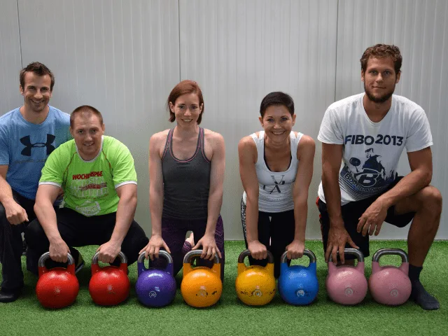 Fünf Personen mit verschiedenfarbigen Kettlebells auf grüner Trainingsmatte, Gruppenfoto