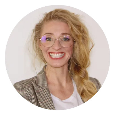 Julia mit blonden Haaren, lila Brille, Blazer und breitem Lächeln
