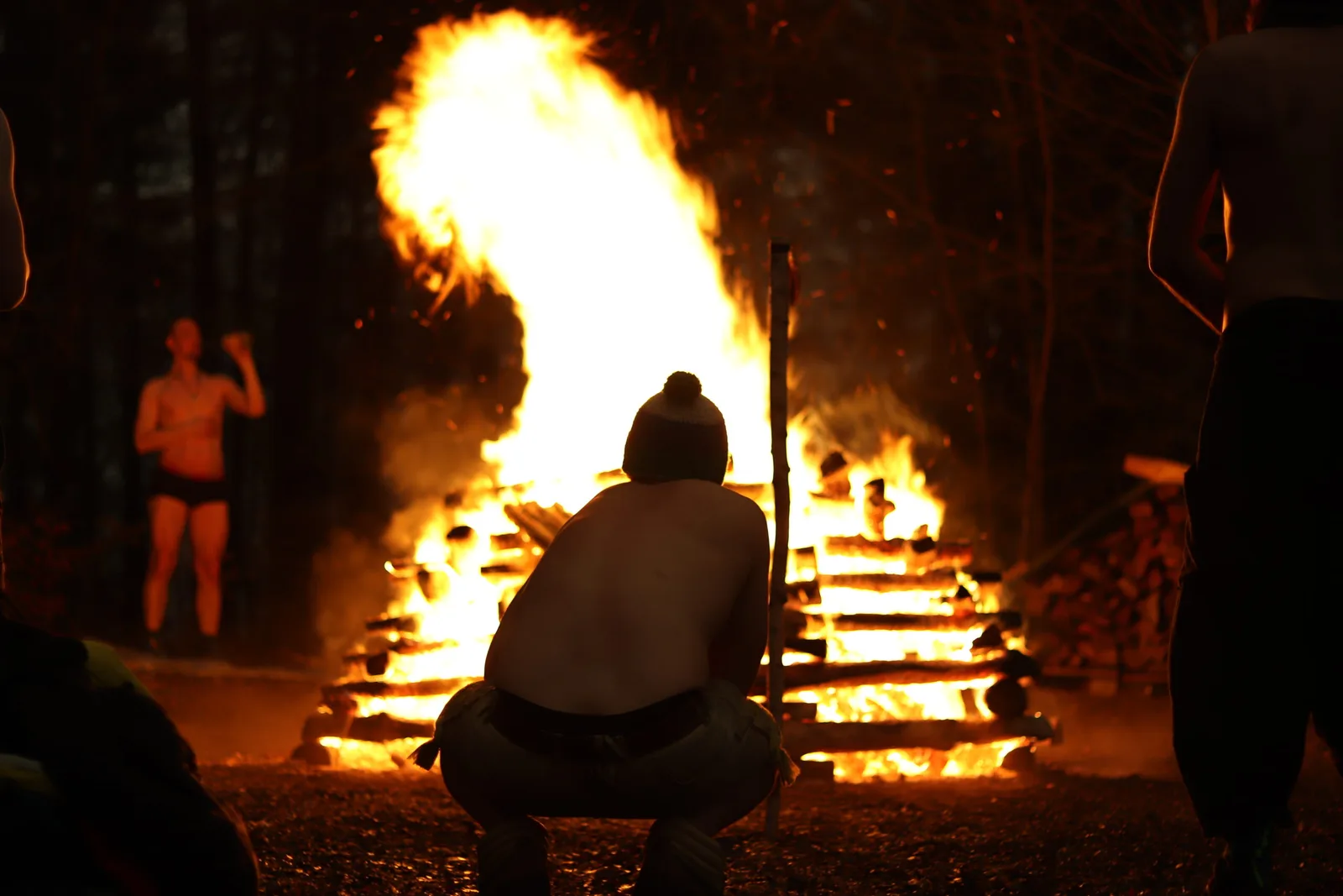 Person sitzt vor großem Lagerfeuer in der Nacht, umgeben von anderen Menschen