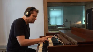 Andreas Czeppel spielt an Keyboard mit Kopfhörern in Musikstudio mit Glasfenster und Aufnahmegerät