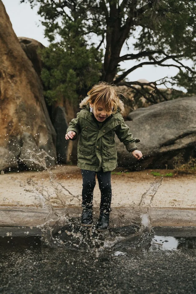 Kind springt lachend in Pfütze, grüne Jacke, Natur mit Bäumen und Steinen