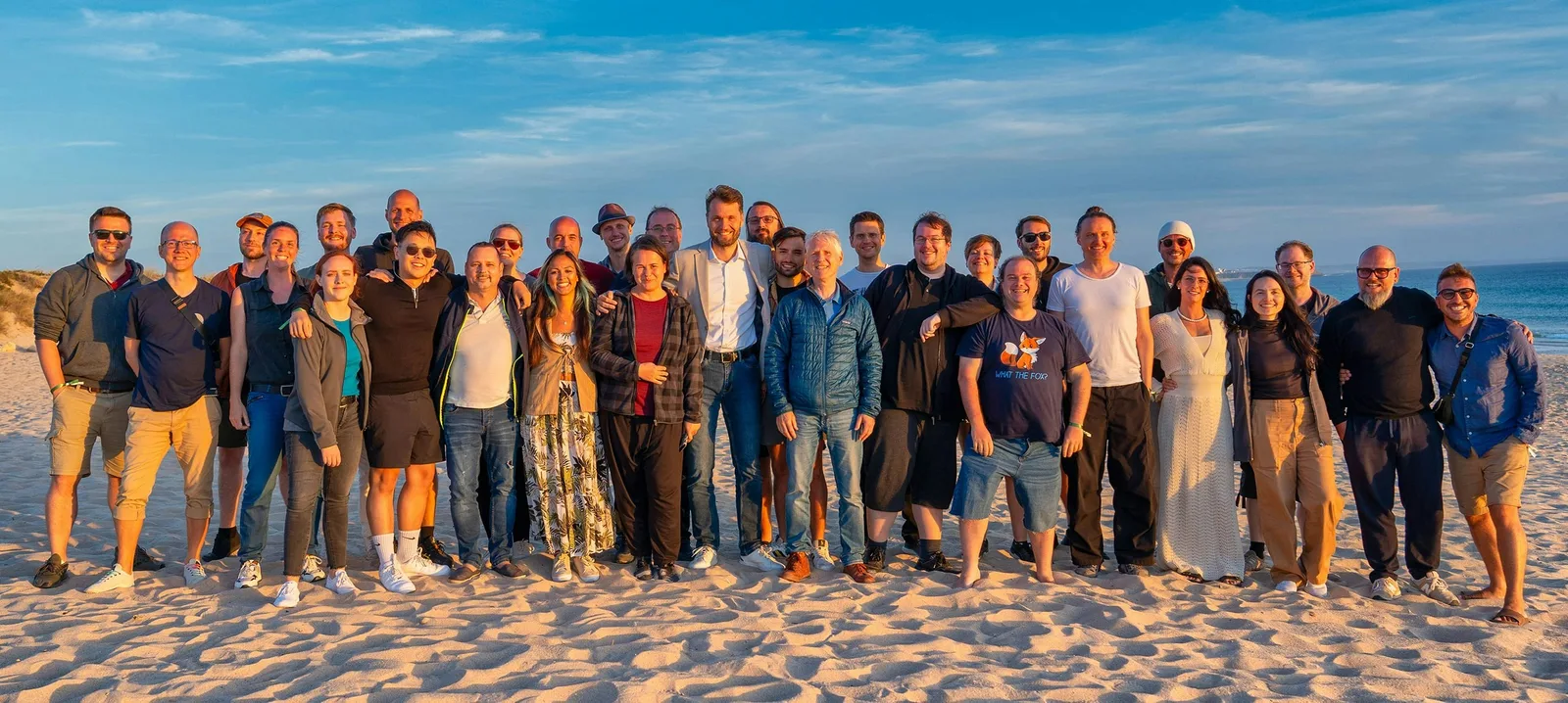 KlickTipp Team am Strand, etwa 30 Personen in lockerer Gruppenformation, blauer Himmel, engagiertes Team hinter erfolgreichen