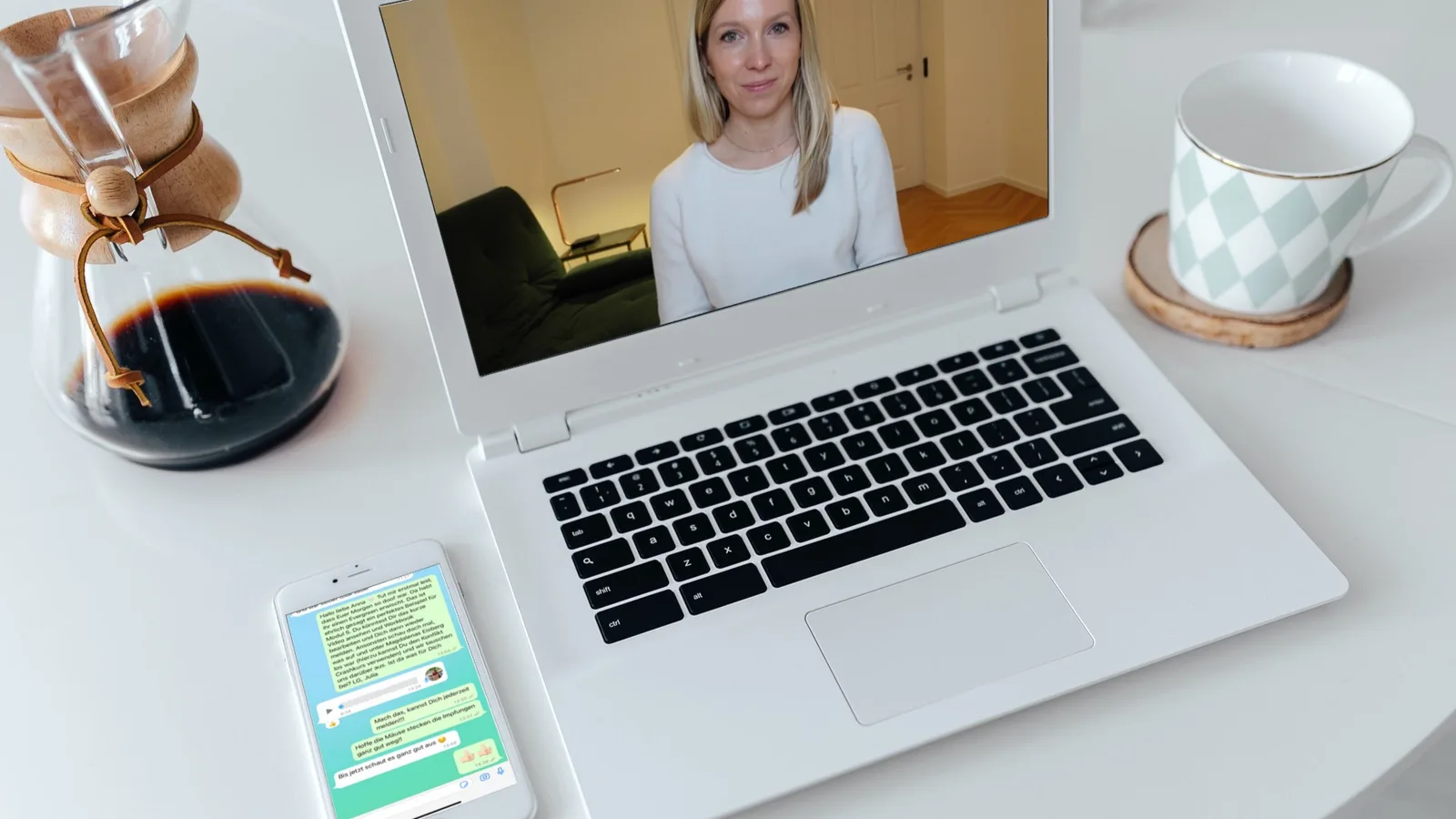 Laptop-Bildschirm zeigt Videokonferenz mit Frau, daneben Kaffeekanne, Tasse, Smartphone mit App-Interface auf weißem Schreibt