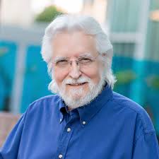 Neale Donald Walsch mit weißem Haar und Bart in blauem Hemd vor städtischem Hintergrund