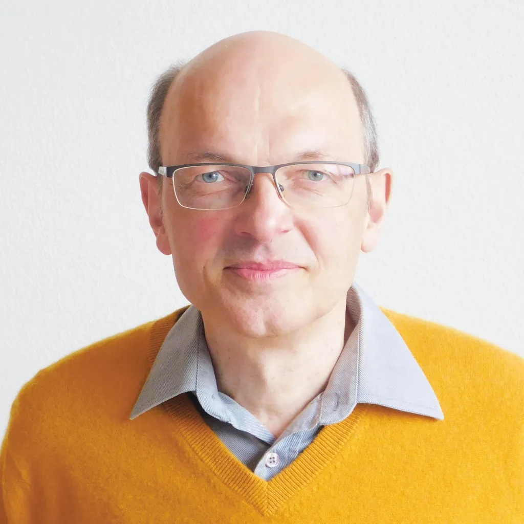 Armin Risi Mann mit Brille in gelbem Pullover und hellblauem Hemdkragen vor hellgrauem Hintergrund
