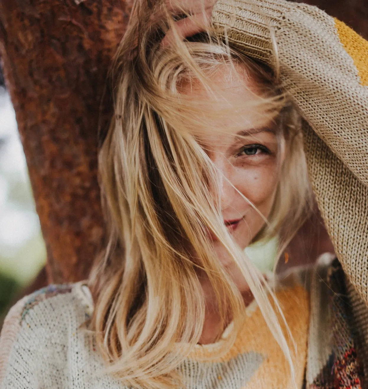 Blonde Frau mit Strickmütze schaut hinter Baumstamm hervor