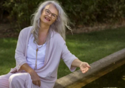 Monika Cardinal sitzt auf Holzbalken im grünen Garten, trägt Brille und lächelt