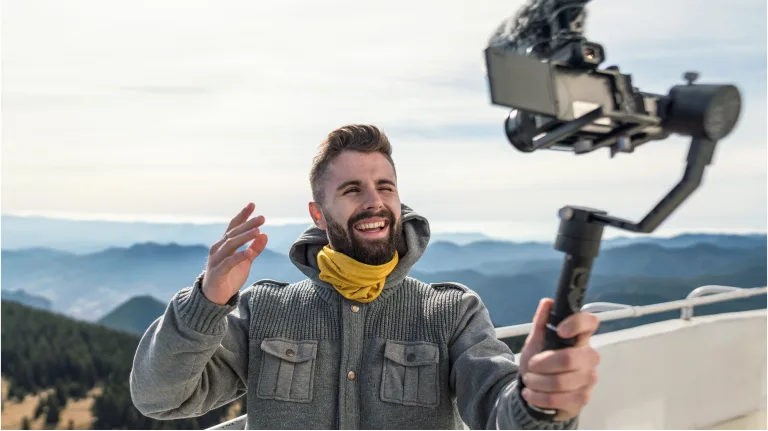 Mann mit Gimbal-Kamera in Berglandschaft, lächelnd und winkend