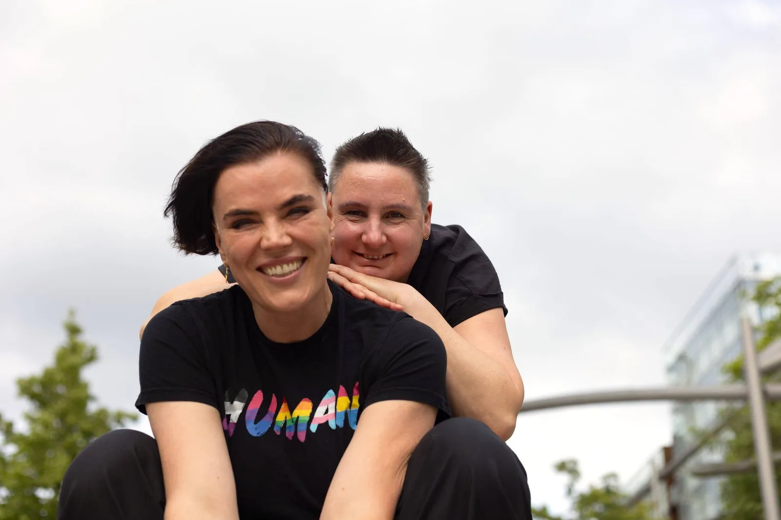 Zwei Frauen umarmen sich lachend vor bewölktem Himmel und Bäumen