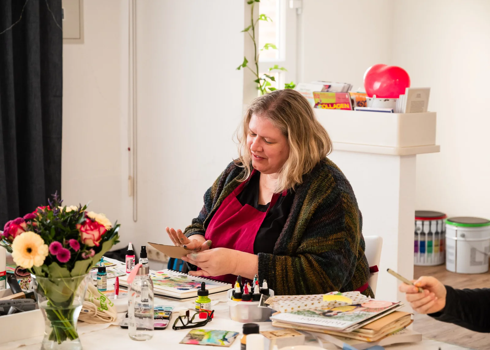 Frau an Kreativtisch mit Kunstmaterialien, Blumen und Kunstwerken arbeitend