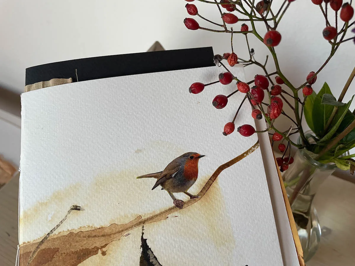 Schwarzes Tagebuch mit Rotkehlchen-Aquarell neben echten roten Beeren auf Holz