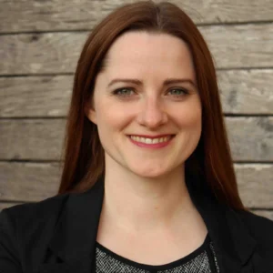 Porträt einer Frau mit rötlich-braunen langen Haaren, schwarzem Blazer und grauem Muster-Top vor Holzhintergrund