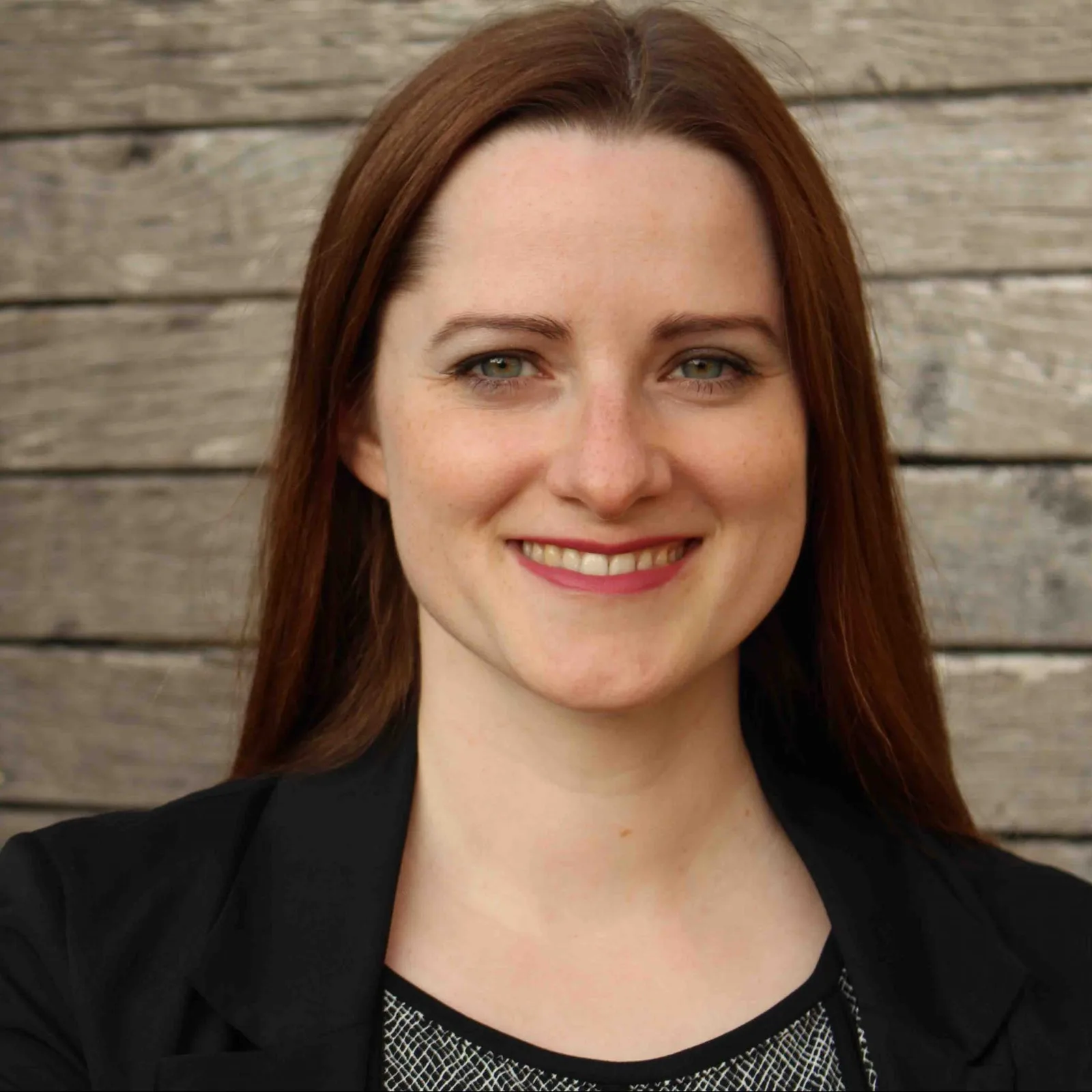 Porträt einer Frau mit rötlich-braunem Haar, schwarzem Blazer und Musteroberteil vor Holzhintergrund