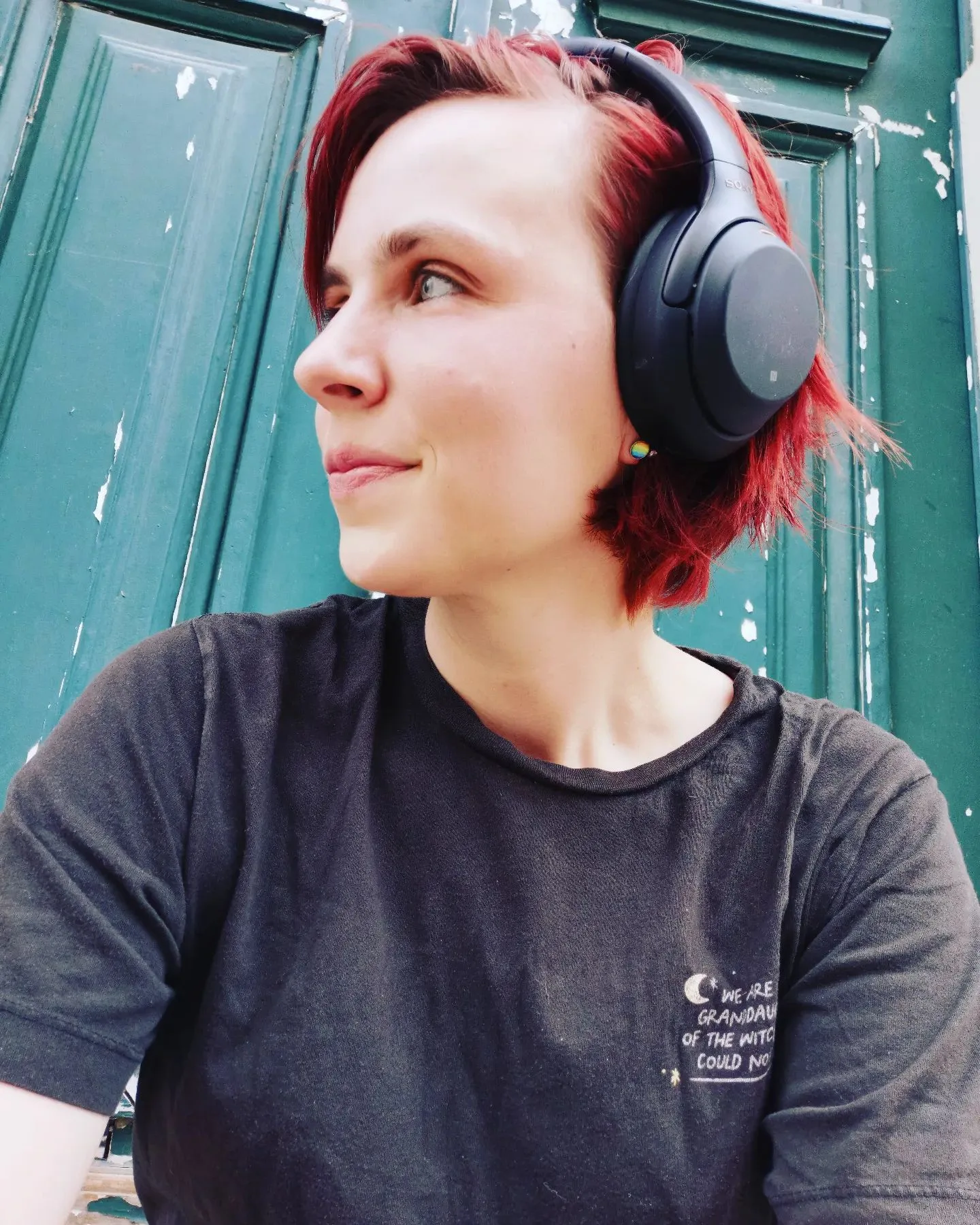 Frau mit rot gefärbten Haaren, Kopfhörer, schwarzes T-Shirt, vor grüner Tür, Seitenansicht