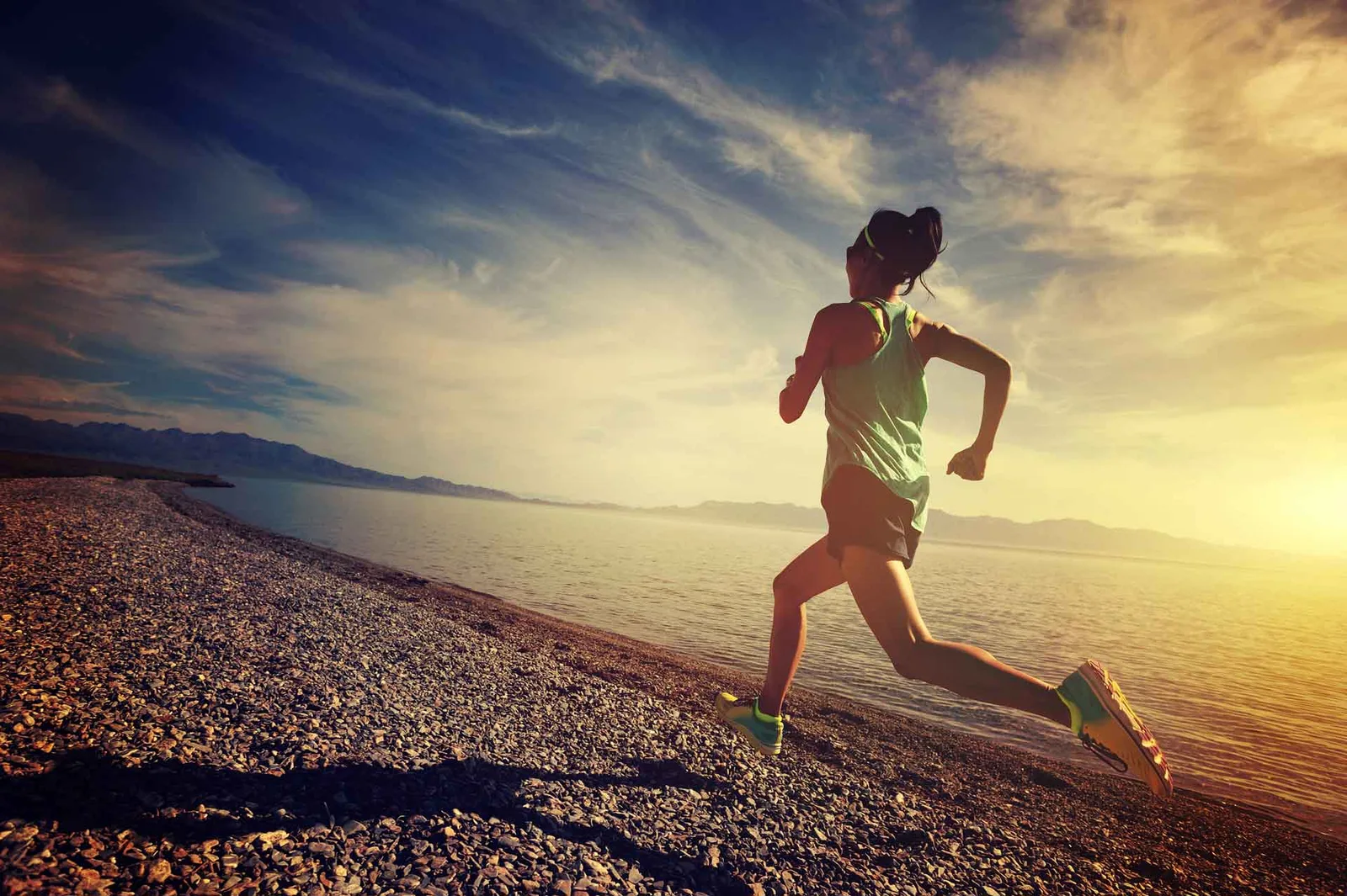 Frau joggt bei Sonnenaufgang am Strand entlang mit Blick aufs Meer und Berglandschaft