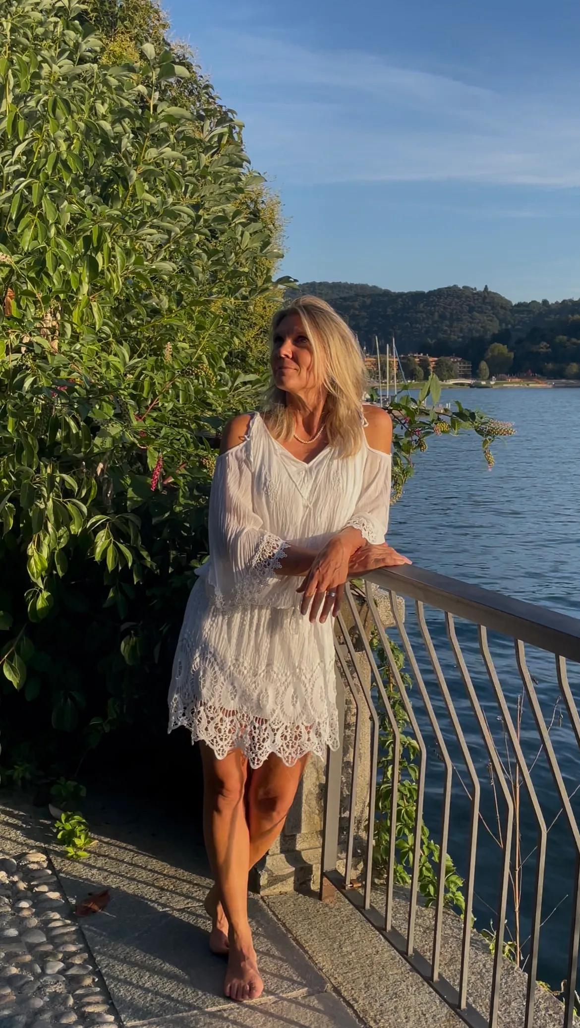 Frau in weißem Kleid an Seeufer mit grünen Pflanzen und Segelbooten