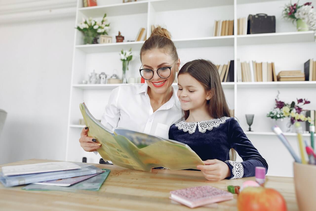 Lerncoach Ausbildung – Zertifikatskurs für Lernbegleitung 4 Frau mit Brille liest mit Schulkind ein Buch am Holztisch in heller Büro-Bibliothek