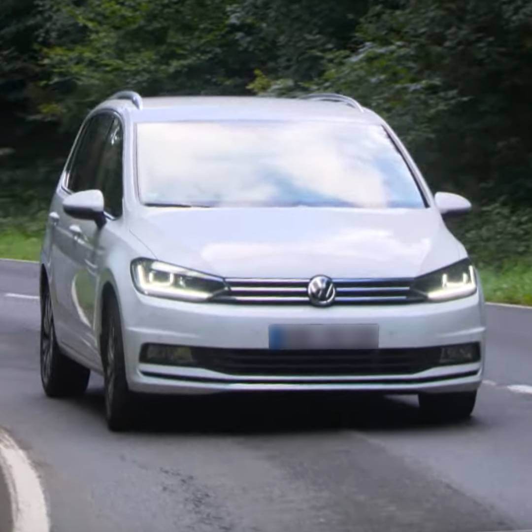 Weißer VW Touran I von vorne auf Straße mit grüner Vegetation im Hintergrund