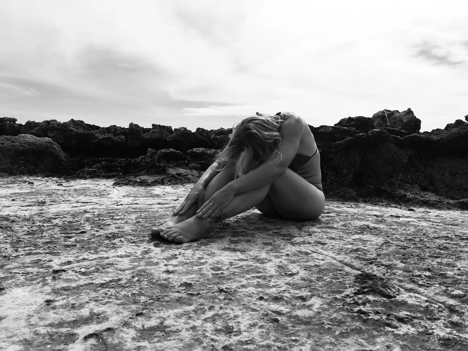 Schwarzweiß-Bild: Frau sitzt am Strand zwischen dunklen Felsen, umarmend