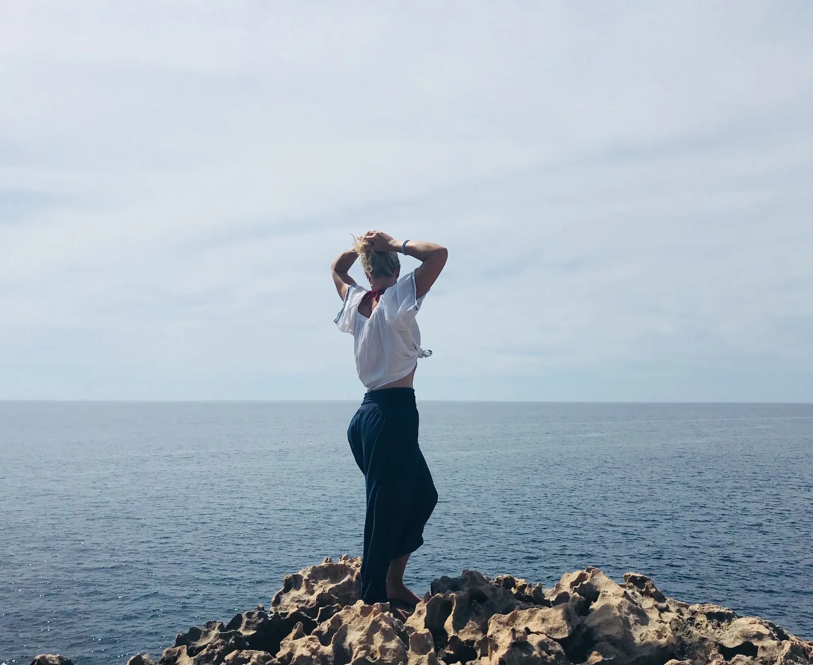 Frau in weißem und dunklem Outfit steht auf Felsen am Meer, Arme hochgesteckt