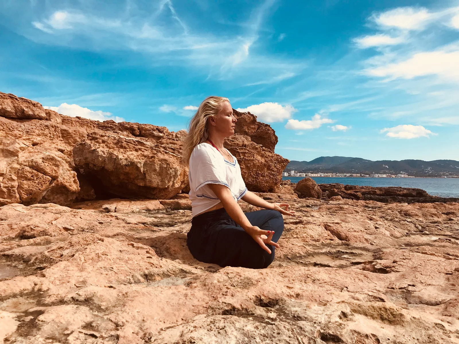 Frau sitzt auf Felsen an Küste mit Blick aufs Meer, trägt weißes T-Shirt