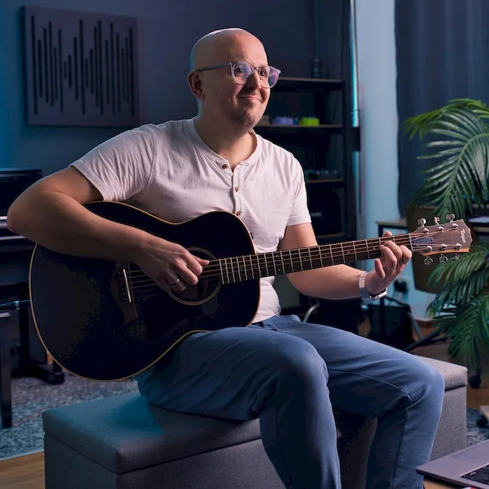 Christian Konrad sitzt mit schwarzer Akustikgitarre in modernem Studio-Setting mit Pflanze, weißes T-Shirt und Brille