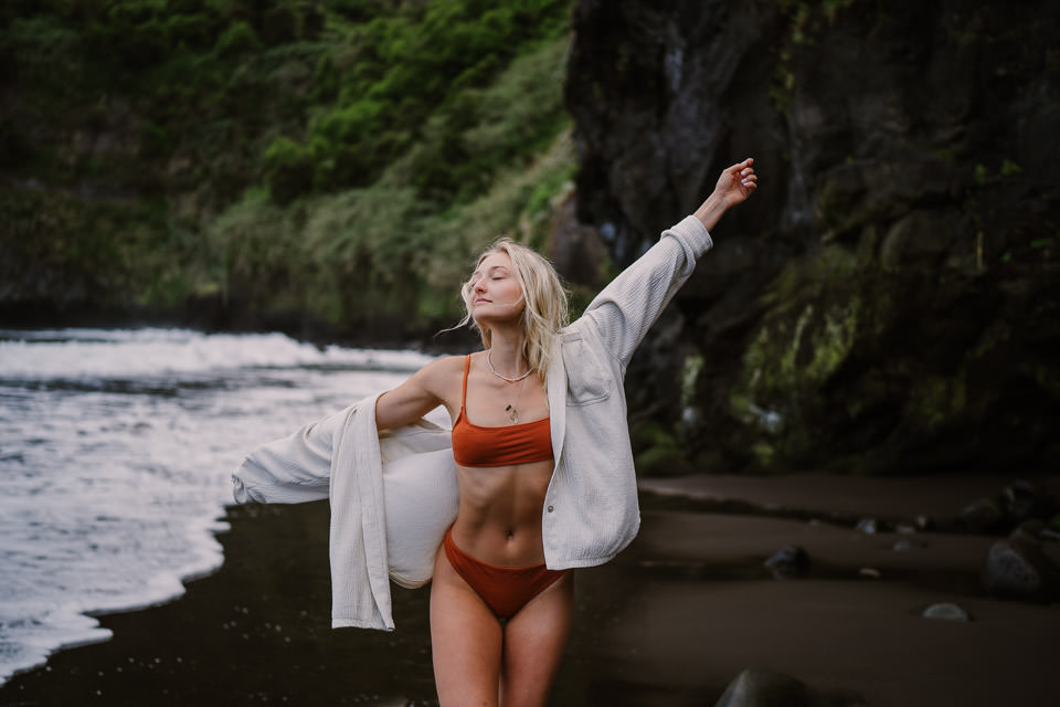 Blondes Mädchen am Strand mit orangem Bikini und weißer Jacke, Felsküste