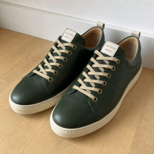 Dunkelgrüner Leder-Sneaker mit Creme-Schnürsenkeln auf hellem Holzboden, Nahaufnahme von oben