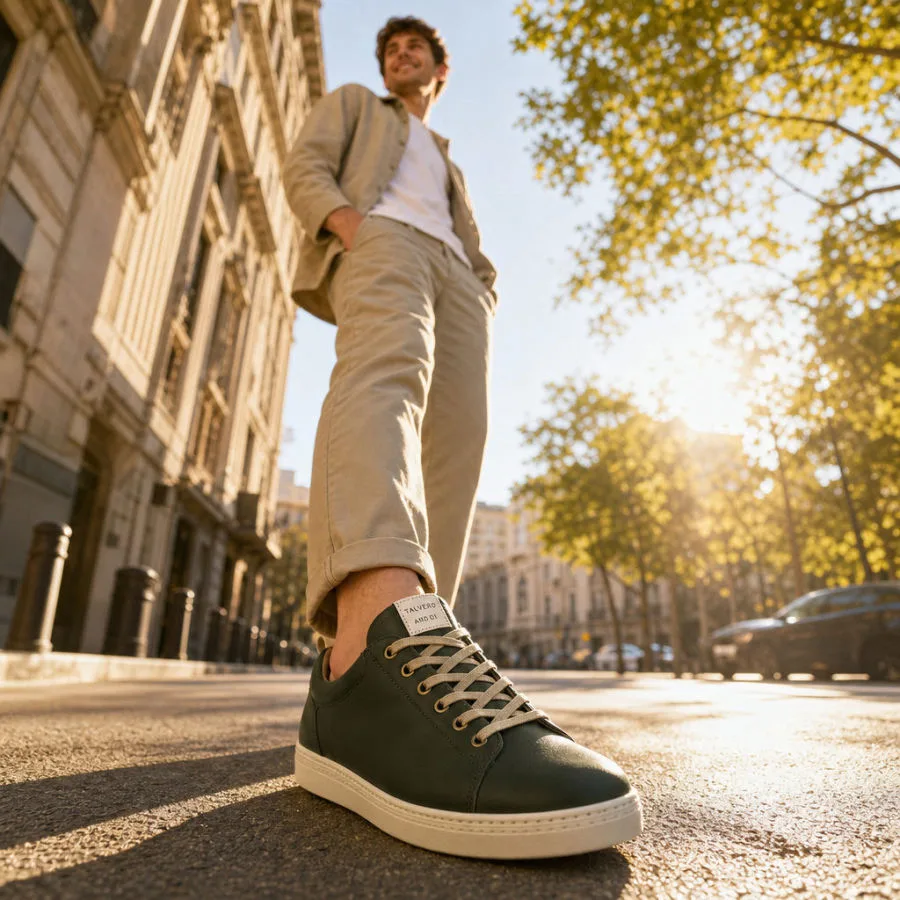Mann trägt dunkelgrüne Sneaker, Unterschenkelaufnahme im urbanen Setting mit Gebäuden und Sonnenlicht