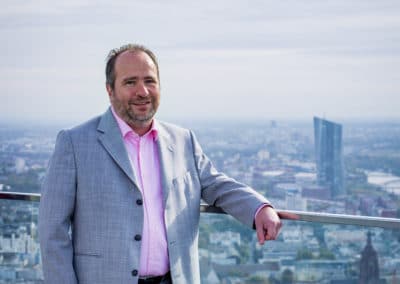Mann in Anzug und rosa Hemd auf Dachterrasse mit Stadtpanorama im Hintergrund