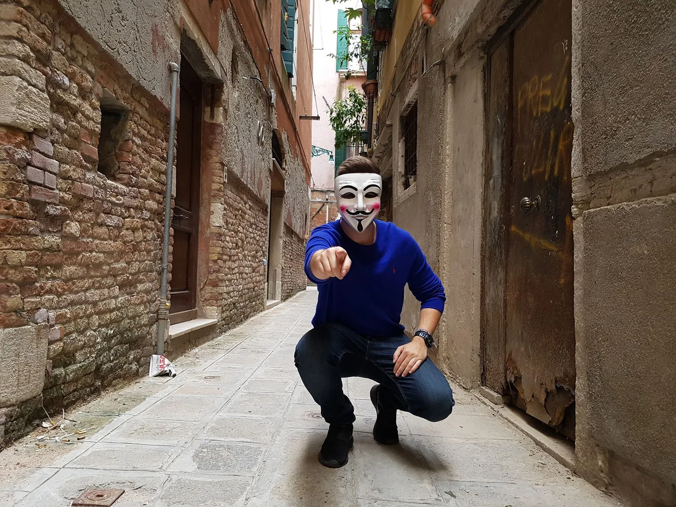 Mann mit Guy-Fawkes-Maske in blauem Pullover zeigt auf Betrachter in Altstadtgasse
