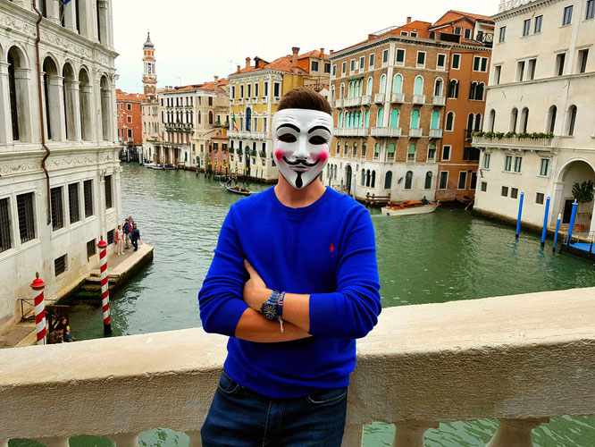 Mann mit Guy-Fawkes-Maske in blauem Pullover posiert vor Venedigs Kanal