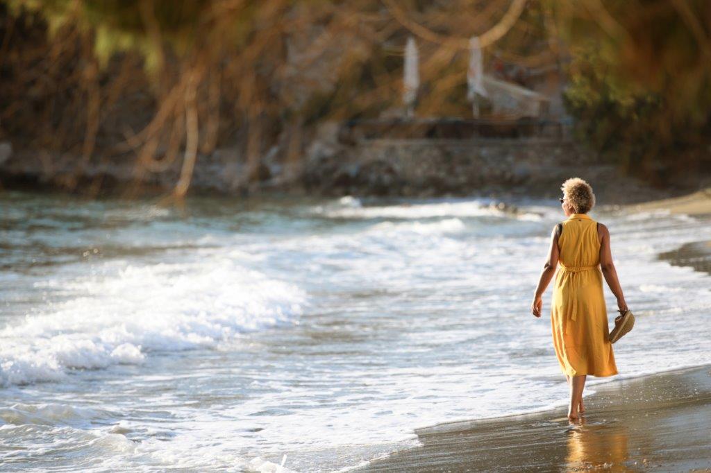 Frau in gelbem Kleid läuft barfuß am Strand entlang mit Wellen im Hintergrund