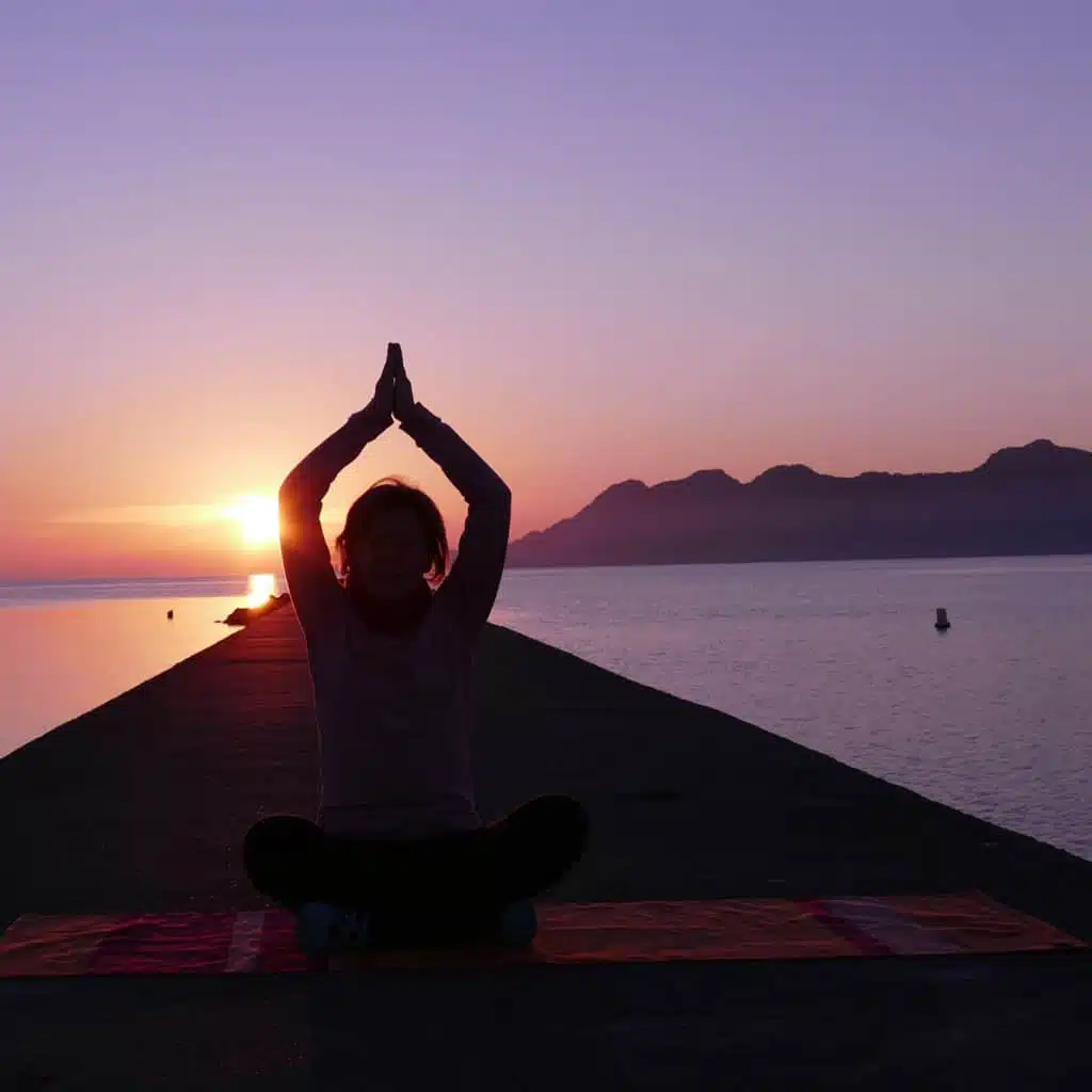 Magische Rauhnächte – Yoga- und Meditationskurs von Yoga mit Martina 4 Frau in Yoga-Pose bei Sonnenaufgang am Meer mit Bergen im Hintergrund, Dankbarkeitspose