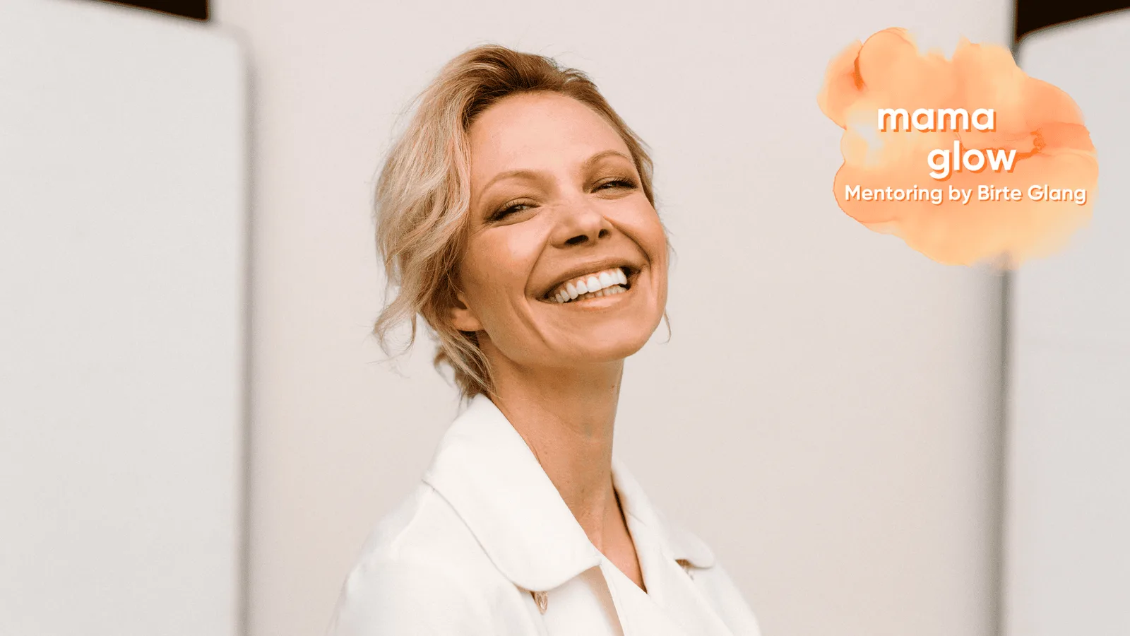 Birte Glang lächelt leuchtend, weißer Blazer, mamaglow Logo orange rechts, Professional