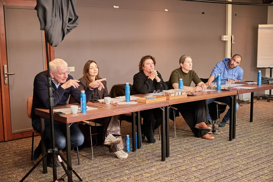 Fünf Personen sitzen an Konferenztisch während Diskussion oder Panel mit Getränken und Unterlagen