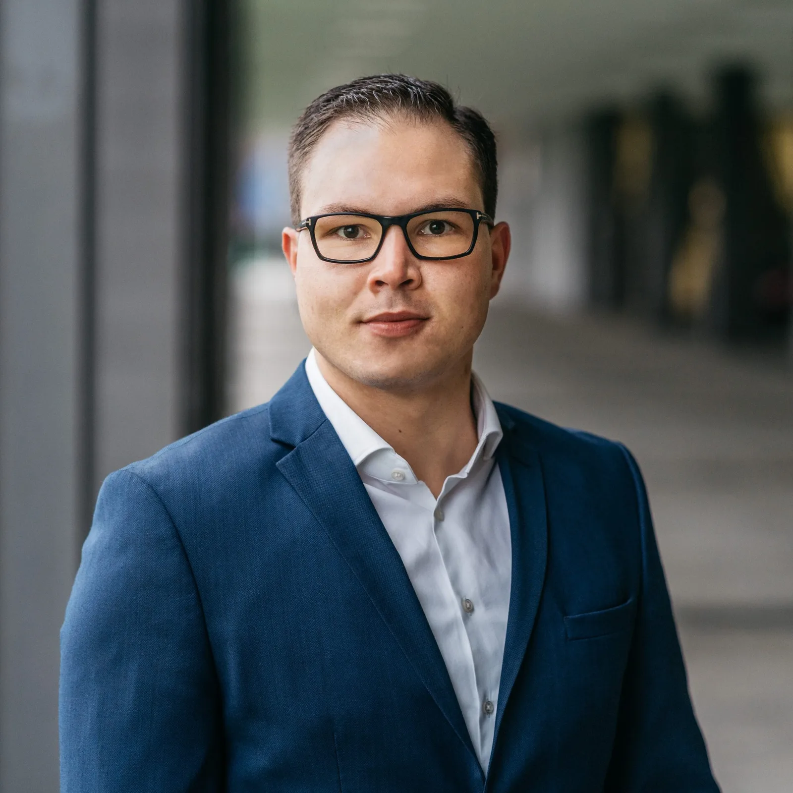 Mechatroniker AP2 – IHK-Prüfungsvorbereitungskurs von Christoph Höller 9 Geschäftsmann mit Brille, dunkelblauen Anzug und weißem Hemd vor unscharfem Bürogebäude-Hintergrund