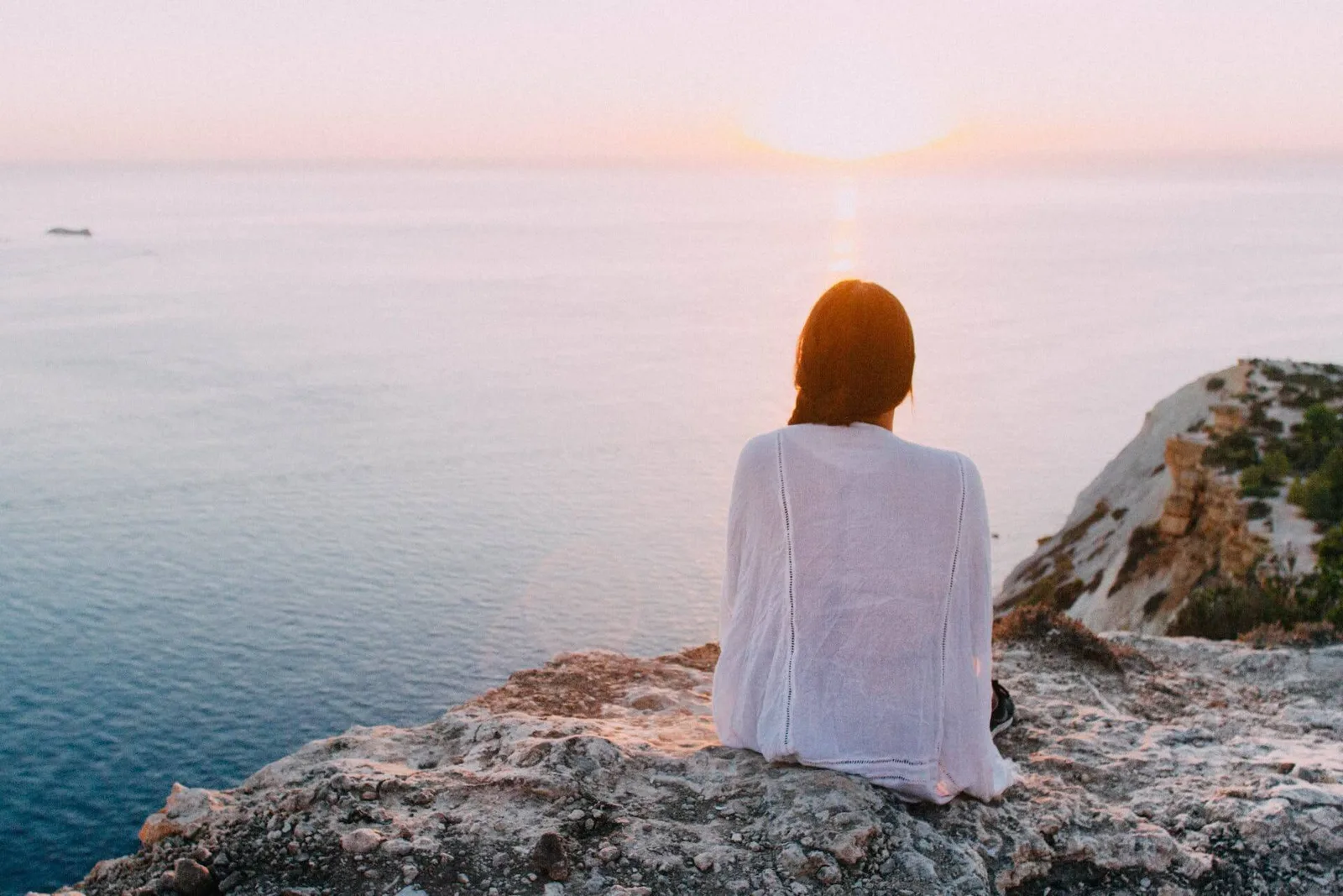 Frau sitzt auf Klippe über Meer bei Sonnenuntergang, Ruhe und innerer Frieden
