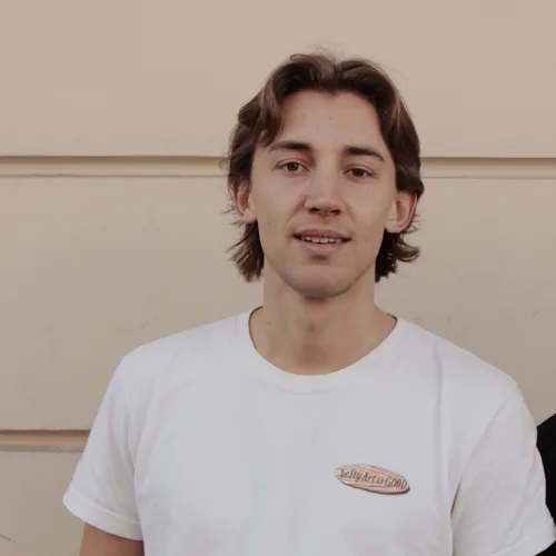 Junger Mann mit braunen Haaren in weißem T-Shirt mit Logo vor beigefarbenem Hintergrund