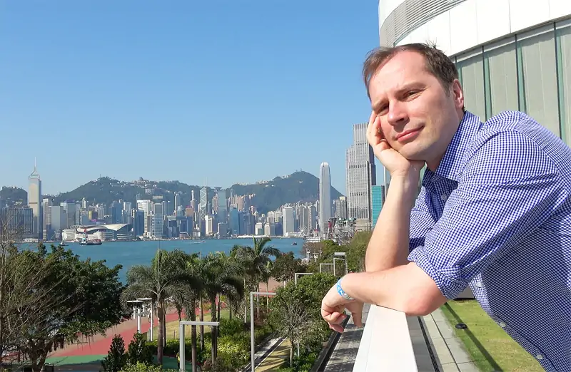 Mann in blaukariertem Hemd lehnt an Balkongeländer mit Hongkong Skyline im Hintergrund