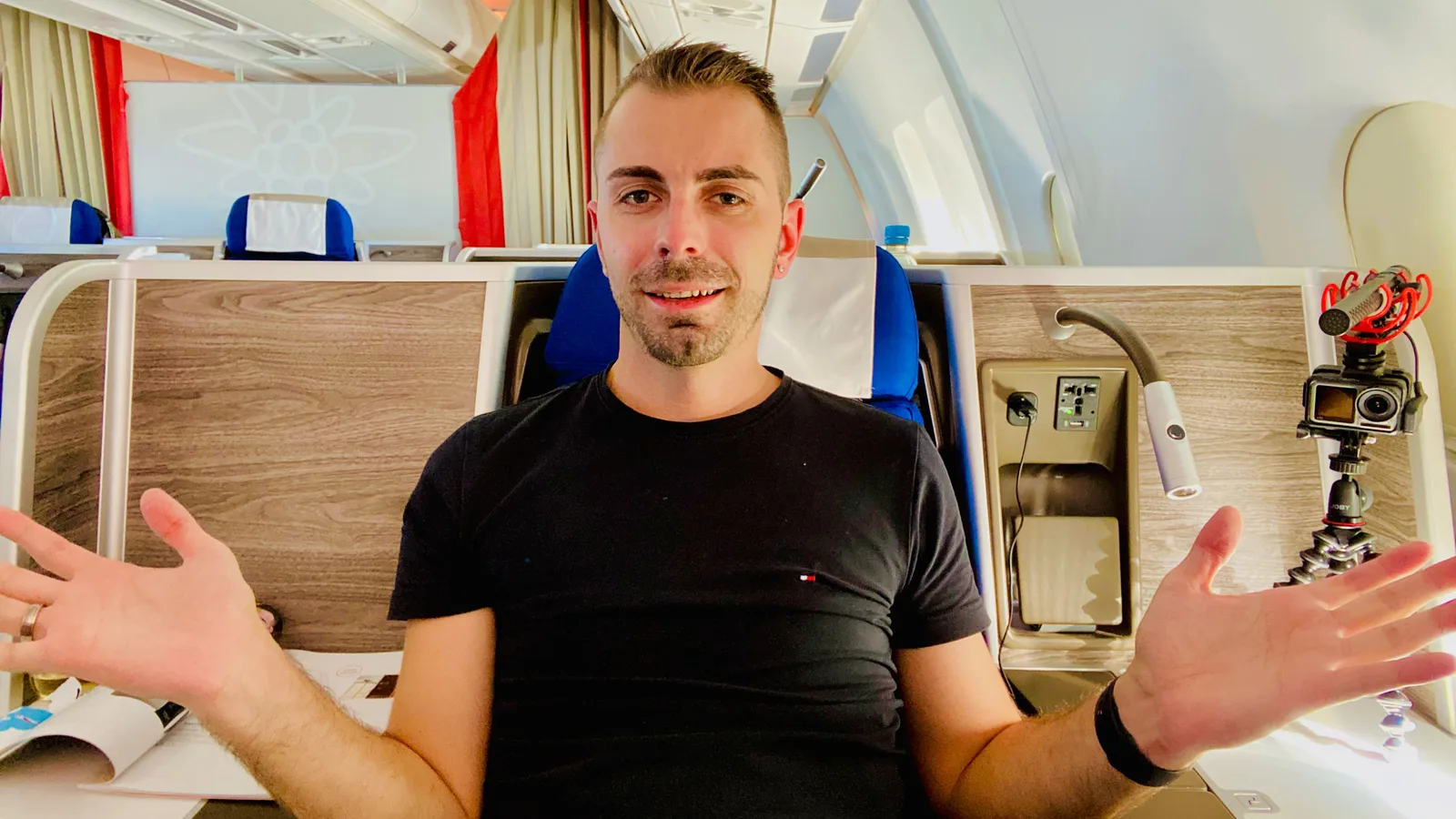 Mann in schwarzem Shirt sitzt in Business-Class-Flugzeugkabine mit roten und blauen Sitzen, spricht in Kamera mit ausgebreite